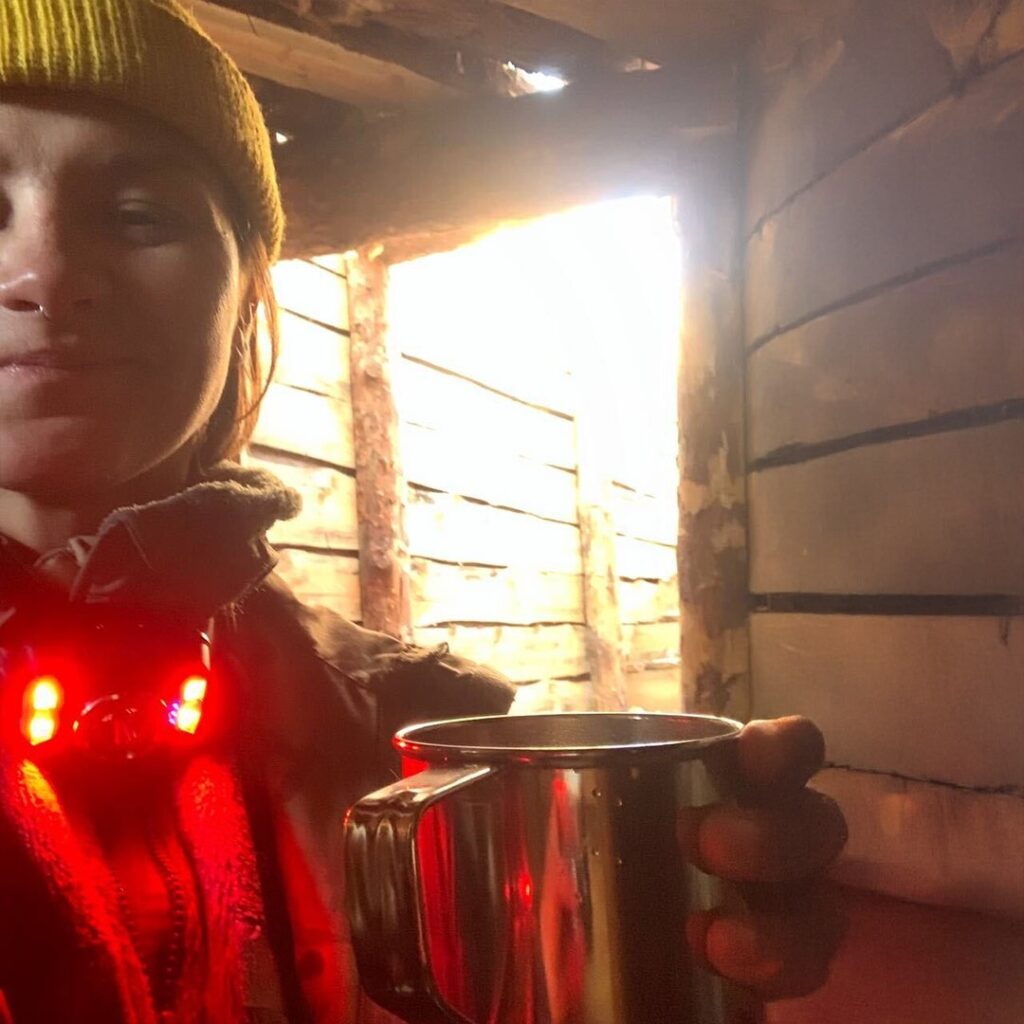 A person in a yellow beanie and outdoor gear holds a metal mug inside a wooden shelter. A faint light shines through the wooden planks, creating a cosy atmosphere.