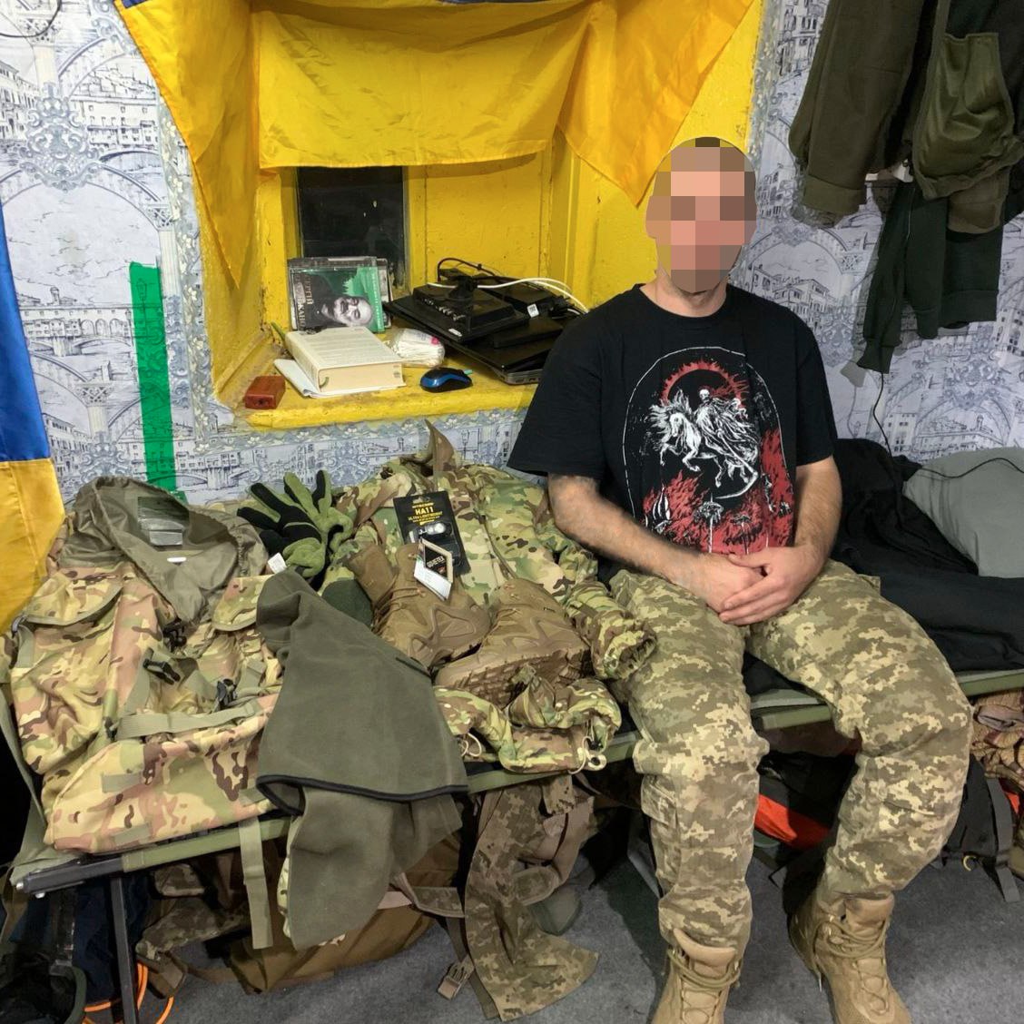 Soldier sitting by a window in a decorated room with military gear laid out on a table next to him.