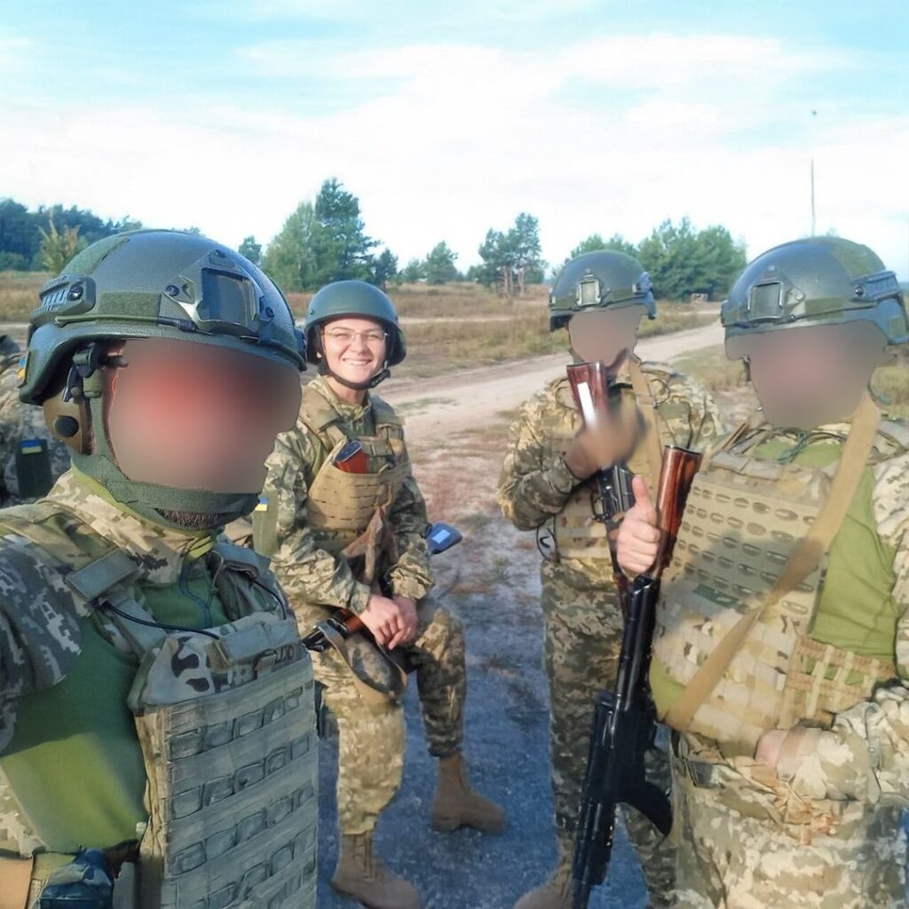 A group of four people in military gear, standing outdoors on a clear day. The central figure is smiling, while the others have their faces blurred.