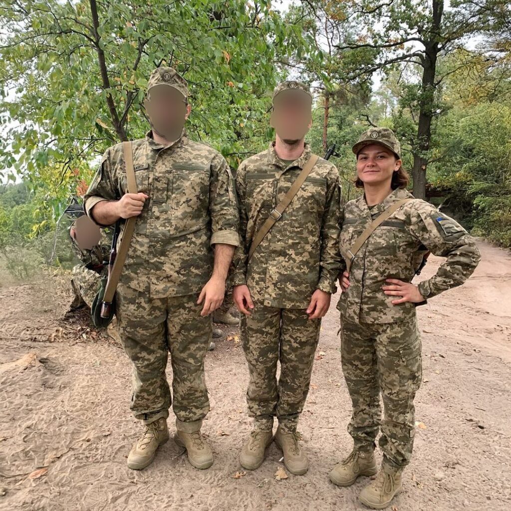 Three individuals in camouflage uniforms pose in a wooded area, with one person smiling confidently with hands on hips. Two faces are blurred, maintaining their anonymity.