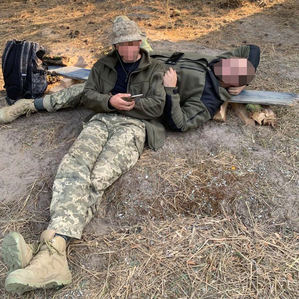 Two soldiers resting on the ground outdoors, one sitting while using a phone, and the other lying down on a makeshift bed.