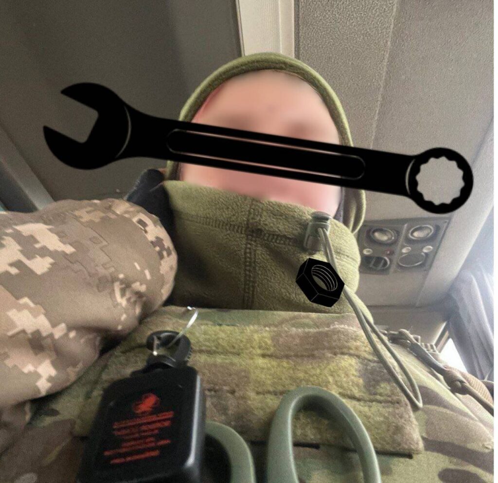 A person in military gear, including a green balaclava and camouflage vest, is pictured on a vehicle. Visible details include a key holder and medical scissors hanging from their vest.