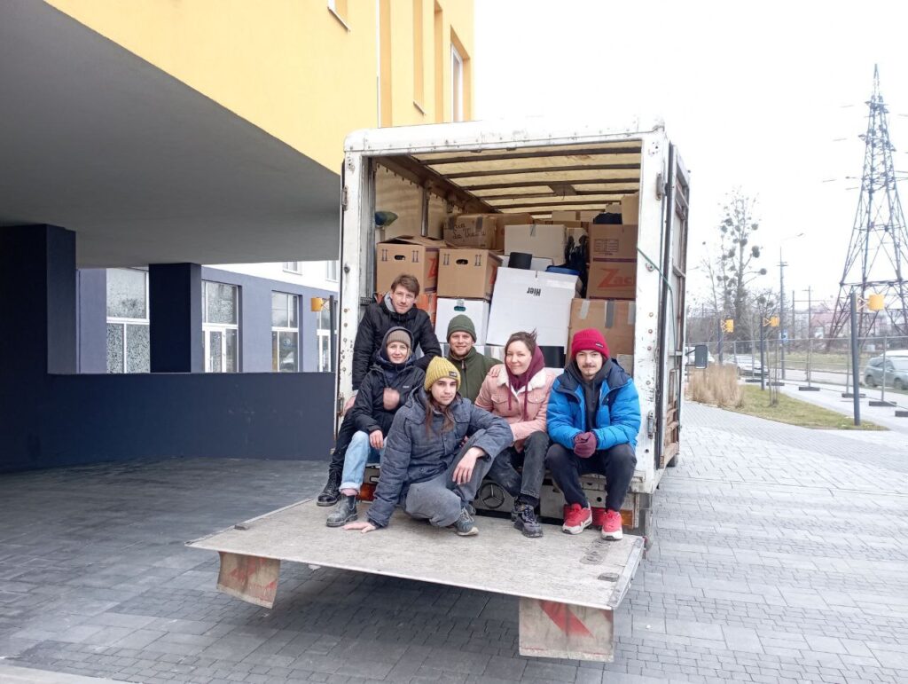 Six people sit on the open lift gate of a white delivery truck filled with boxes labeled “Zack.” They are outdoors near a modern building with power lines in the background.