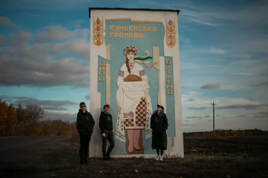 Three people standing next to a painted welcome sign for the Kunyev hromada, showing a traditional Ukrainian woman holding bread against a bright blue sky.