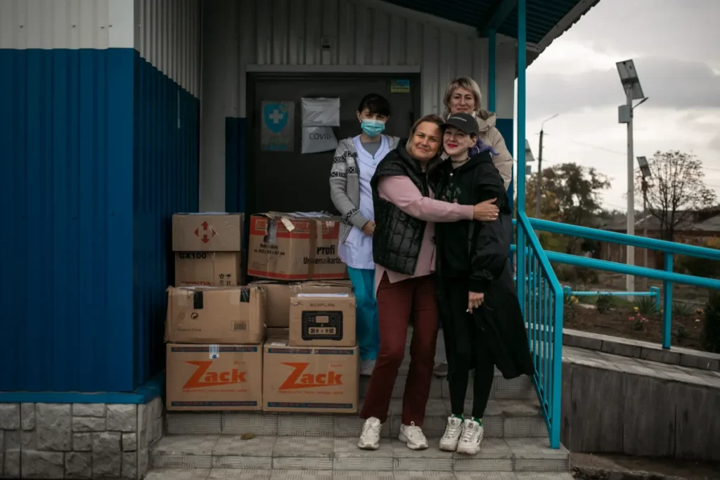 A group of women standing outside a building with boxes of humanitarian aid supplies stacked on the ground near them, smiling and embracing.