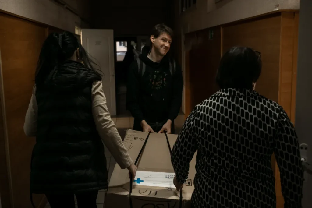 A man smiling as he carries a large box, accompanied by two women, down a corridor of a building.