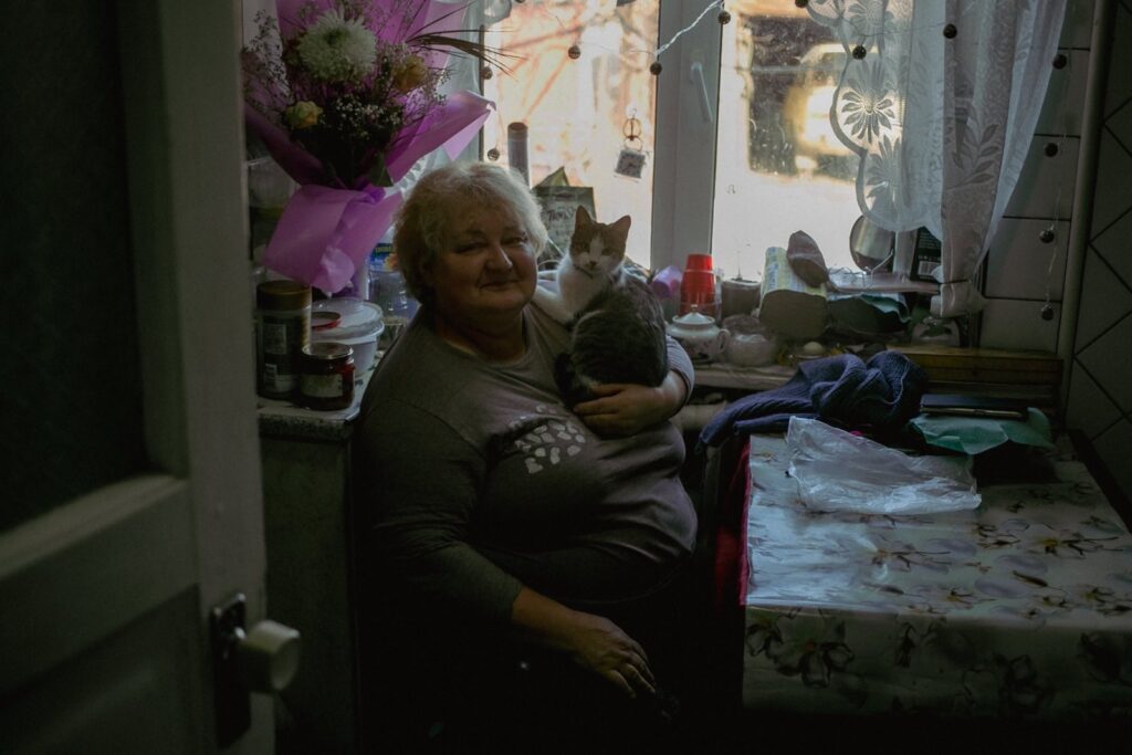 An elderly woman with short, curly white hair sits in a cozy kitchen, holding a grey and white cat close to her chest. The table is covered with a floral tablecloth, and a bouquet of flowers in pink wrapping is placed near the window.