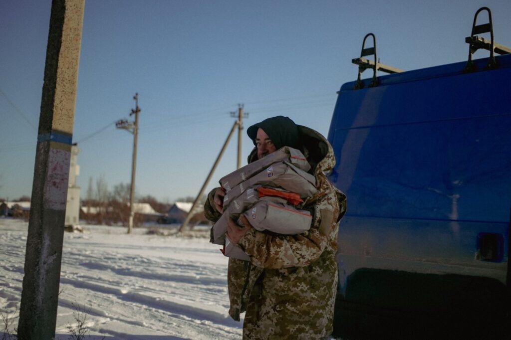 A person wearing a camouflage jacket and hood stands in a snowy outdoor setting, holding multiple packages wrapped in plastic. A blue van is parked behind them, with utility poles and a building visible in the distance.