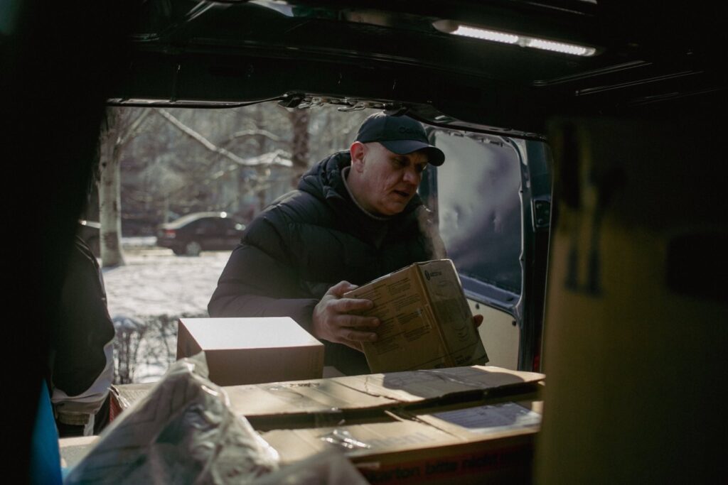 A man in a black puffer jacket and cap unloads cardboard boxes from the back of a vehicle. Steam is visible in the cold air, and the setting is a snowy urban environment with trees and cars in the background.