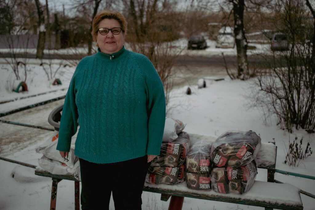 A woman in a teal sweater stands outside in the snow with several bags of pet food stacked on a bench beside her. The background includes trees, a road, and parked cars.