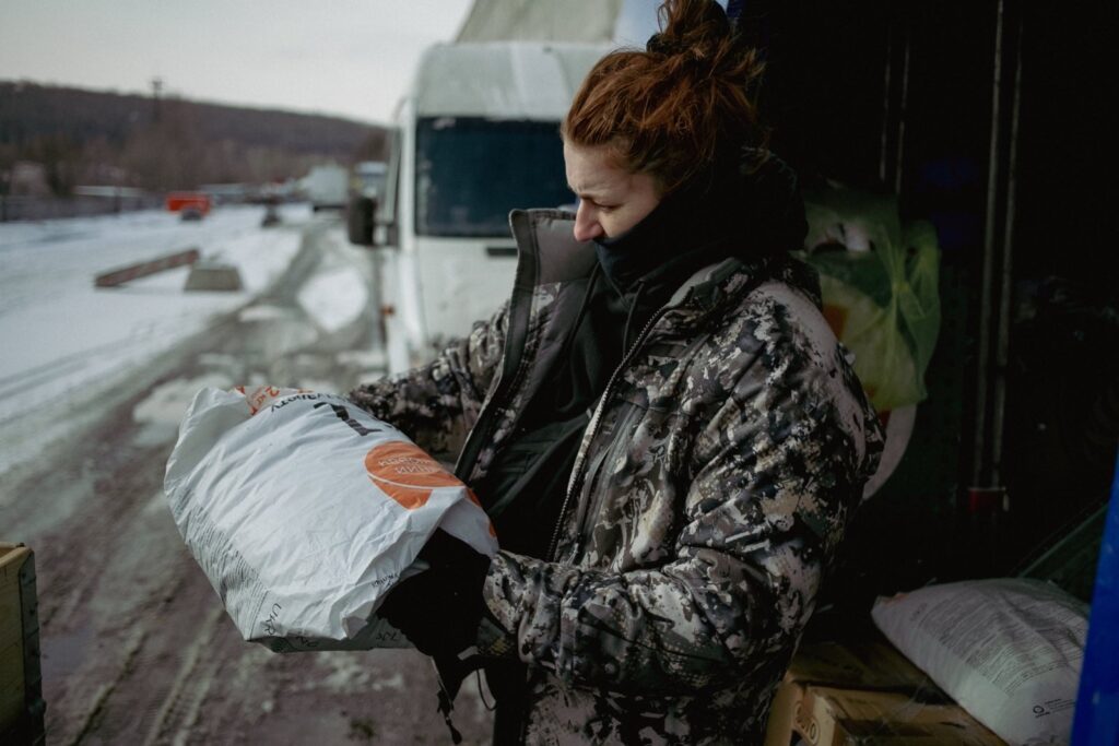 A woman in a camouflage winter jacket examines a large bag of supplies near an open van. A snowy road with multiple trucks and vehicles is visible in the background.