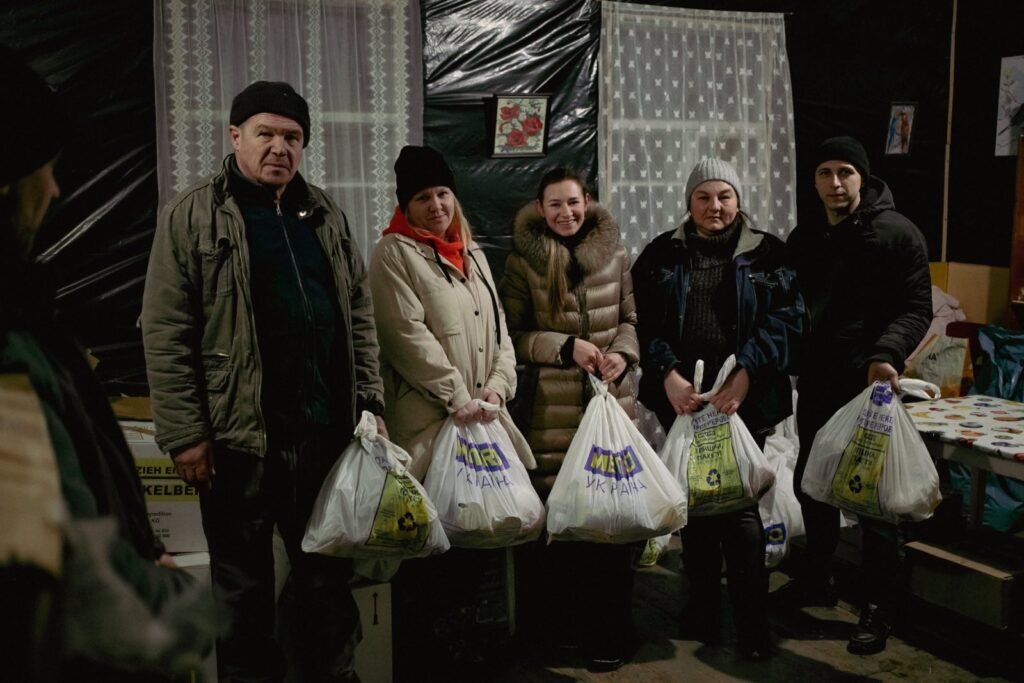 A group of five people stands together indoors, holding bags of humanitarian aid labeled “METRO Україна.” The setting is a makeshift relief distribution center with plastic-covered walls.