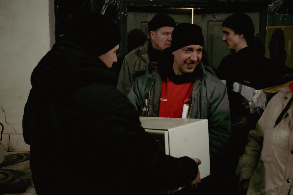 A group of volunteers in winter clothing, including beanies and jackets, work together to unload supplies. A man in a red shirt and green jacket is smiling while assisting with the distribution.