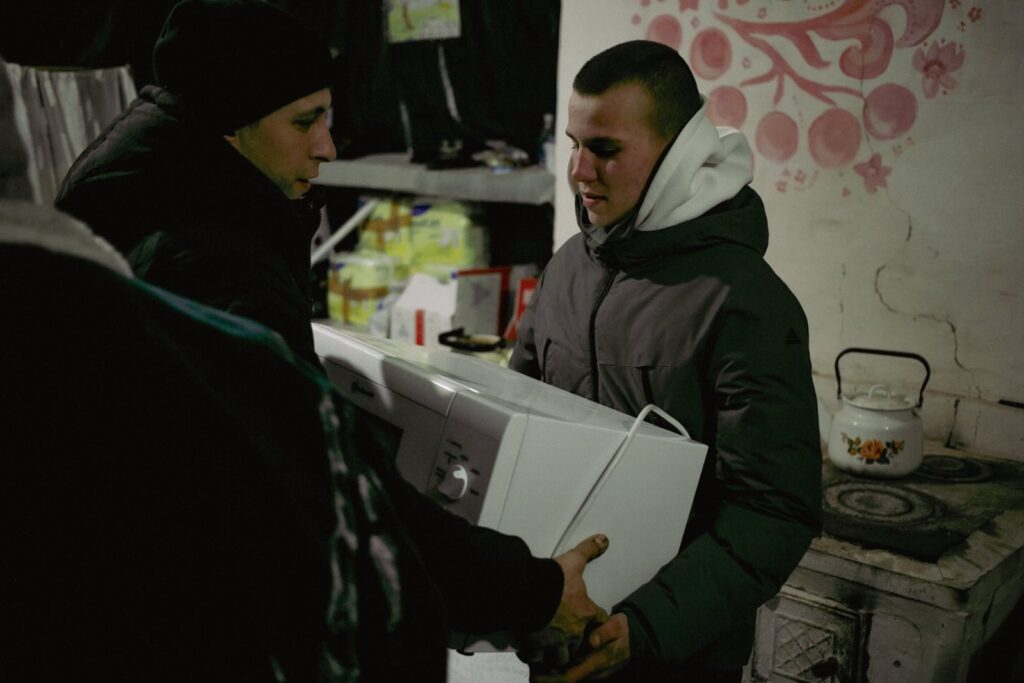 Two men in winter jackets pass a microwave to each other inside a relief center with a rustic interior. A traditional stove with a floral design is visible in the background.